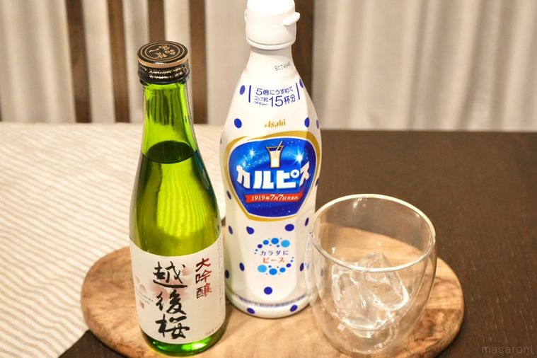 木のカッティングボードにのった日本酒の緑色の瓶とカルピスの原液ボトルと氷入りのグラスとストライプの布