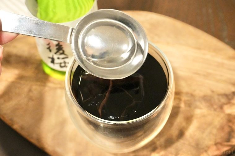 木のカッティングボードにのった日本酒の緑色の瓶と氷とアイスコーヒー入りのグラスに軽量スプーンで日本酒を加える様子