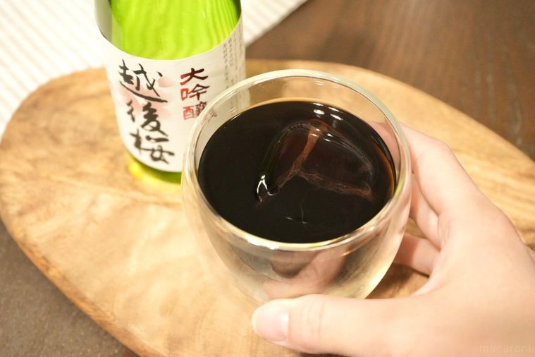 木のカッティングボードにのった日本酒の緑色の瓶と氷とアイスコーヒー入りのグラスを手に持つ様子