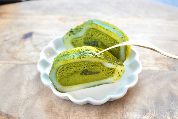 木のカッティングボードと白い小皿にのった緑色の抹茶大福の断面と金のフォークとグレーの布