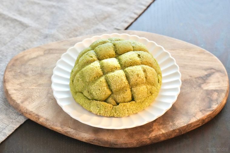 木のカッティングボードと白い皿にのった緑色の抹茶とあんこのメロンパンとグレーの布