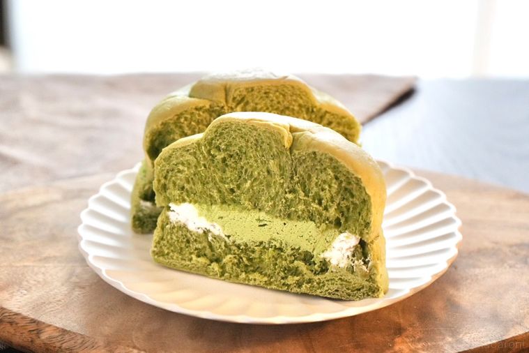 木のカッティングボードと白い皿にのった抹茶とミルクホイップクリームサンドの菓子パンの断面とグレーの布