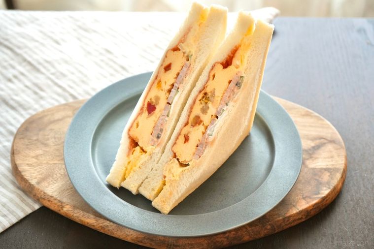 木のカッティングボードとグレーの皿にのったオムレツ入りのサンドイッチとストライプの布