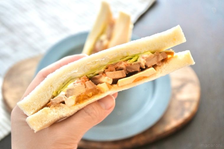 木のカッティングボードとグレーの皿にのった照り焼きチキン入りのサンドイッチを持つ手とストライプの布