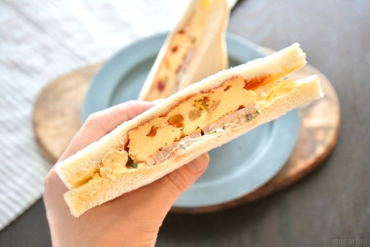 木のカッティングボードとグレーの皿にのったオムレツ入りのサンドイッチを持つ手とストライプの布