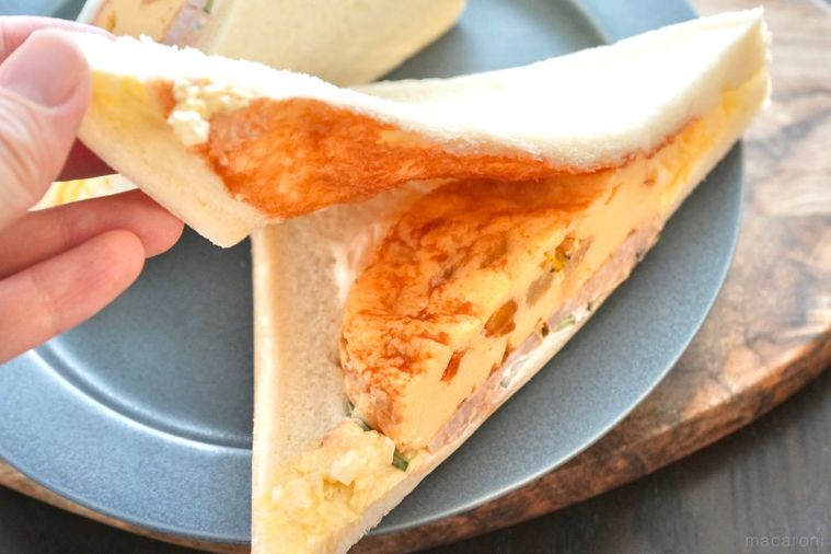 木のカッティングボードとグレーの皿にのったオムレツ入りのサンドイッチをめくる様子とストライプの布