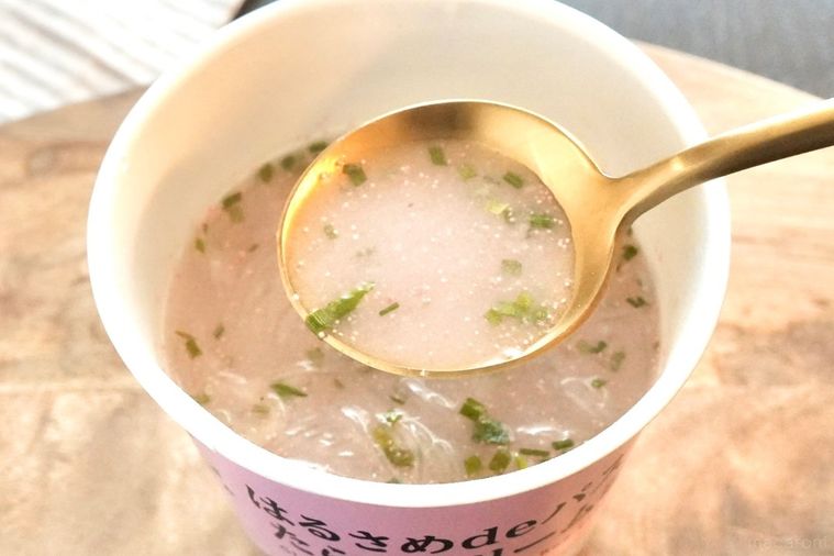 木のカッティングボードにのったピンク色のカップはるさめパスタたらこクリーム味と金と黒のスプーンでスープをすくう様子