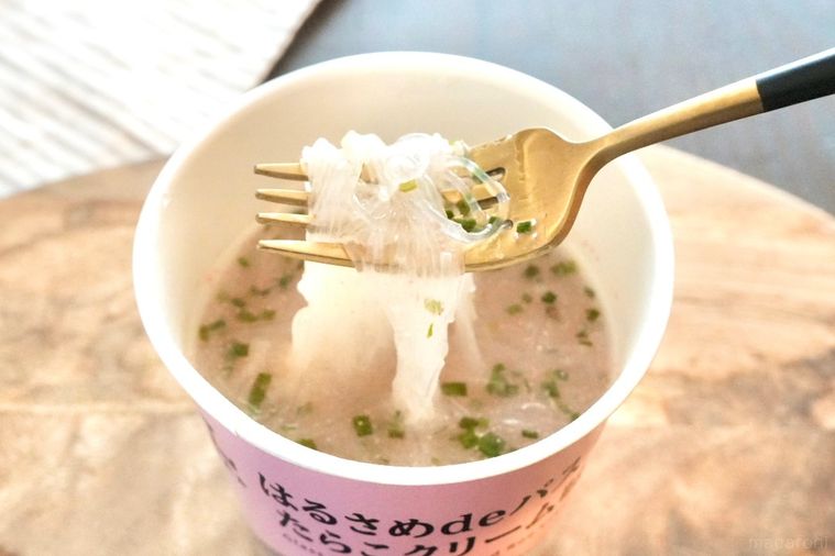 木のカッティングボードにのったピン色のカップはるさめパスタたらこクリーム味を金と黒のフォークで食べる様子とストライプの布