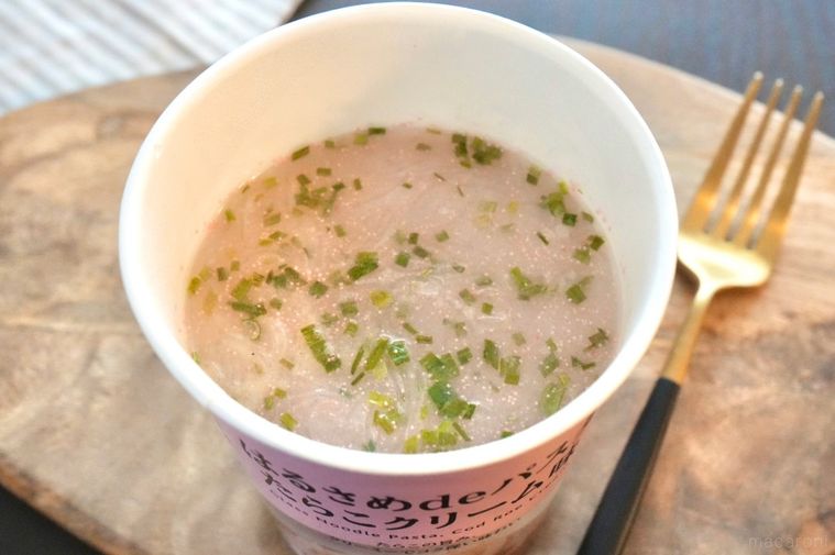 木のカッティングボードにのったピンク色のカップはるさめパスタたらこクリーム味と金と黒のフォークとストライプの布