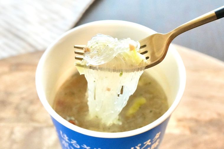 木のカッティングボードにのった青色のカップはるさめパスタボンゴレビアンコ味を金と黒のフォークで食べる様子とストライプの布