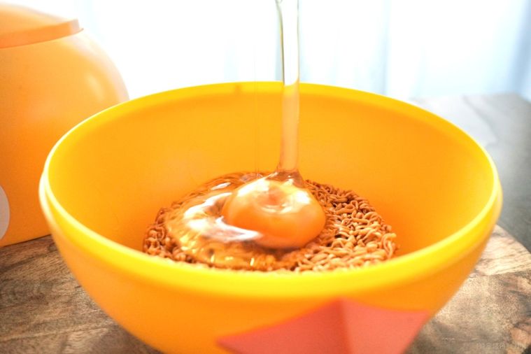 黄色いひよこ型のどんぶりのチキンラーメンにたまごを割って落とす様子