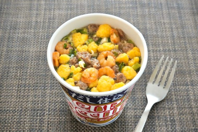 茶色いランチョンマットと和風な黄金色パッケージのだし仕込みカップヌードルと銀色のフォーク