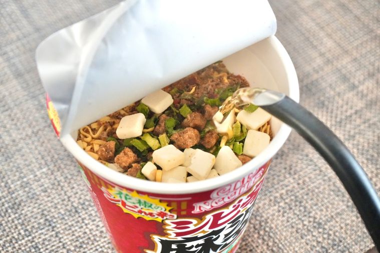 茶色いランチョンマットにのった赤色のパッケージのカップヌードル花椒香るシビ辛麻婆ビッグに黒いポットで熱湯を注ぐ様子