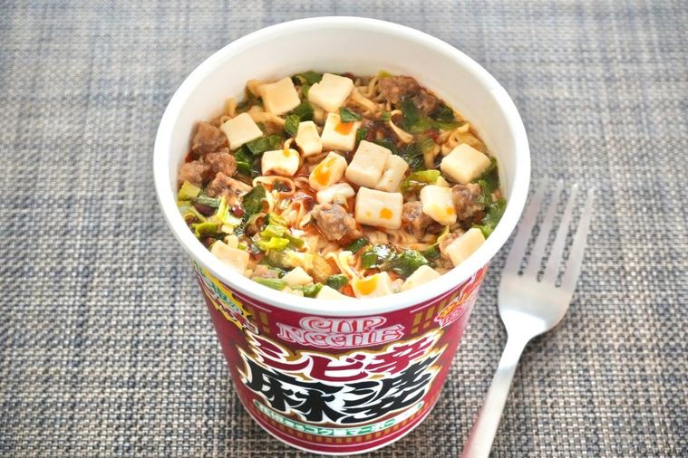 茶色いランチョンマットにのった赤色のパッケージのカップヌードル花椒香るシビ辛麻婆ビッグと銀色のフォーク