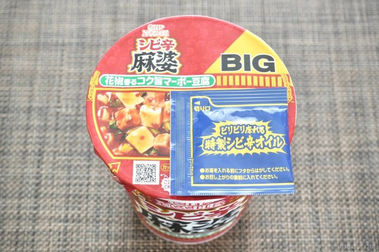 茶色いランチョンマットにのった赤色のパッケージのカップヌードル花椒香るシビ辛麻婆ビッグと青い別袋の特製シビ辛オイル