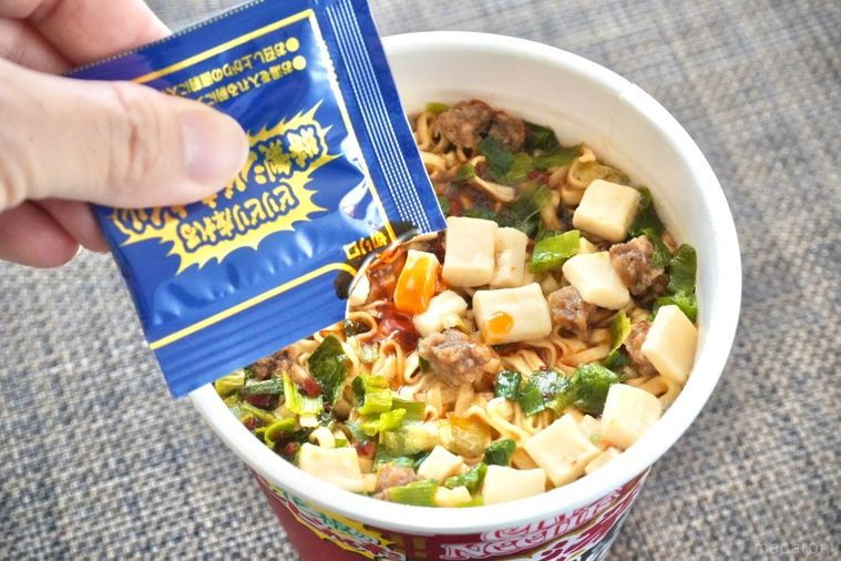 茶色いランチョンマットにのった赤色のパッケージのカップヌードル花椒香るシビ辛麻婆ビッグに青い袋調味油をトッピングする様子