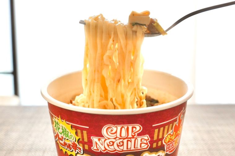 茶色いランチョンマットにのった赤色のパッケージのカップヌードル花椒香るシビ辛麻婆ビッグを銀のフォークで持ち上げる様子