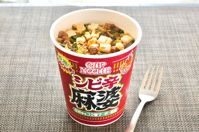 茶色いランチョンマットにのった赤色のパッケージのカップヌードル花椒香るシビ辛麻婆ビッグと銀のフォーク