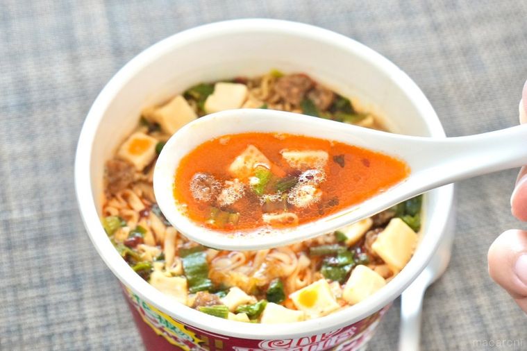 茶色いランチョンマットにのった赤色のパッケージのカップヌードル花椒香るシビ辛麻婆ビッグのスープを白いレンゲですくう様子