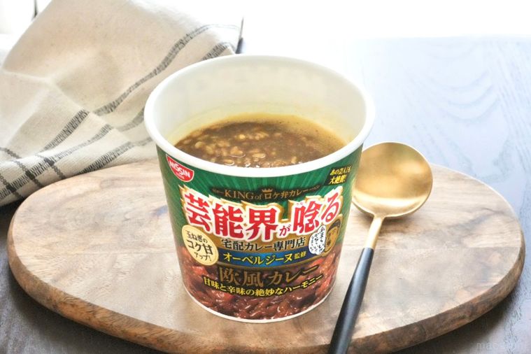 木のカッティングボードにのった緑色のパッケージのカレーカップメシと黒と金色のスプーンとベージュの布