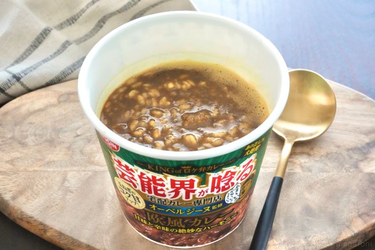 木のカッティングボードにのった緑色のパッケージのカレーカップメシと黒と金色のスプーンとベージュの布