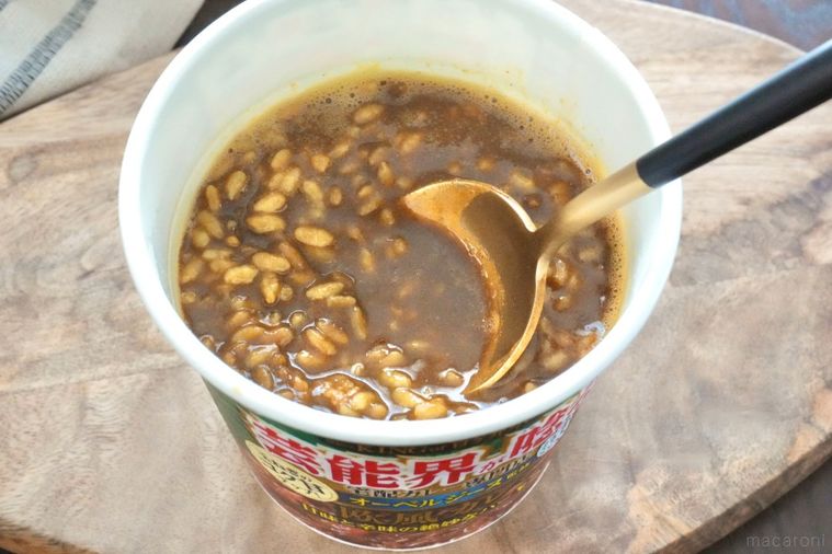 木のカッティングボードにのった緑色のパッケージのカレーカップメシを黒と金色のスプーンですくう様子