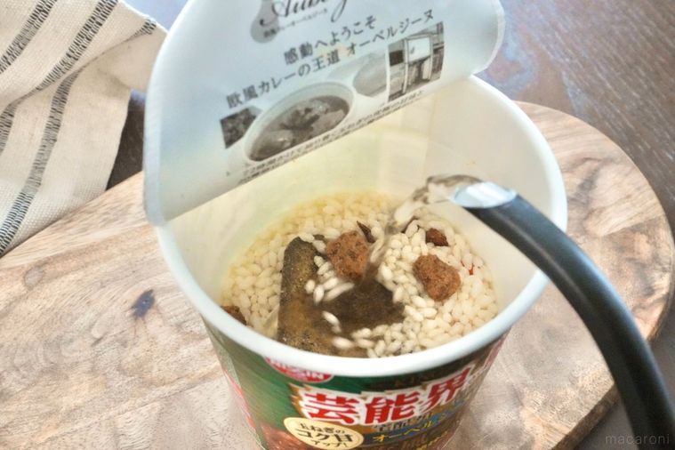 木のカッティングボードにのったカレーカップメシに黒いポットで熱湯を注ぐ様子とベージュの布
