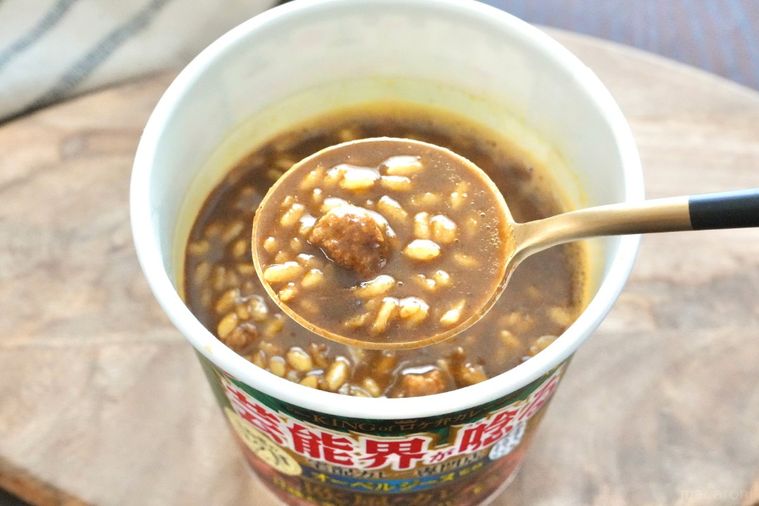 木のカッティングボードにのった緑色のパッケージのカレーカップメシを黒と金色のスプーンですくう様子