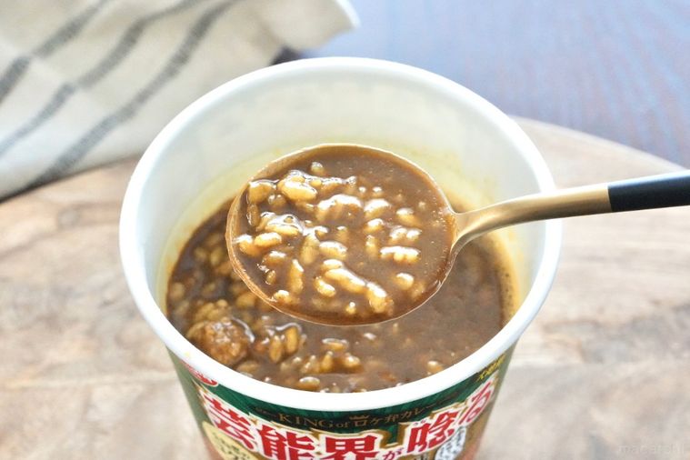 木のカッティングボードにのった緑色のパッケージのカレーカップメシを黒と金色のスプーンですくう様子