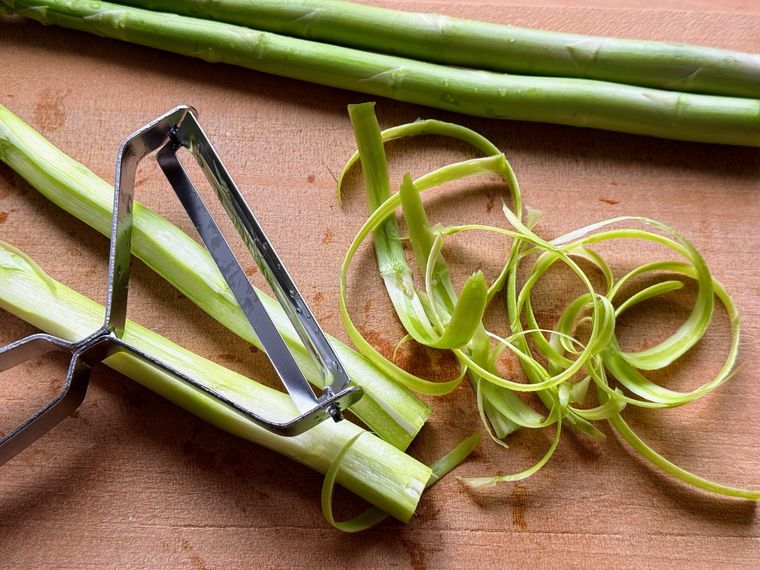 木のカッティングボードにアスパラ、ピーラー、アスパラの皮がのっている様子