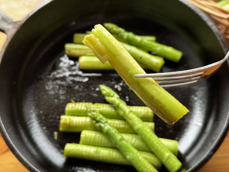 フォークに刺さったアスパラと奥に鉄のフライパンとアスパラソテーが見えている様子