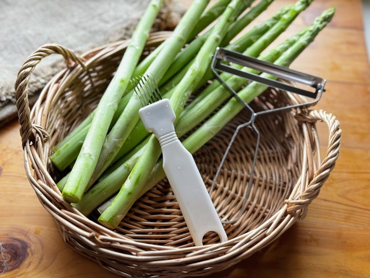 丸いかごに入った複数のアスパラガスと100均のネギカッターとピーラー