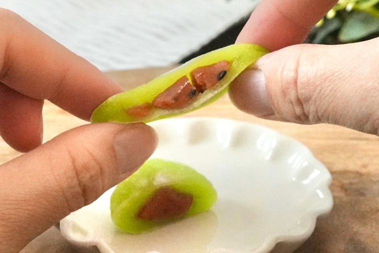 木のカッティングボードと白い皿にのった緑色のすいか味の大福を引き伸ばす手とパッケージとストライプの布