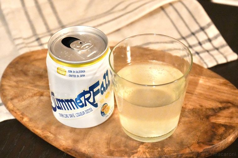 木のカッティングボードにのった日本酒スパークリングの白と黄色のパッケージ缶とグラスと生成色の布