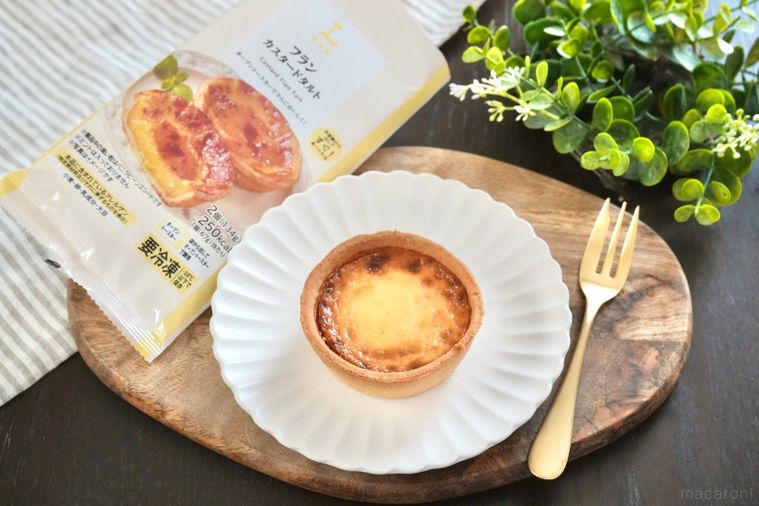 木のカッティングボードと白い皿にのったカスタードタルトとパッケージと金のフォークとストライプの布とグリーン