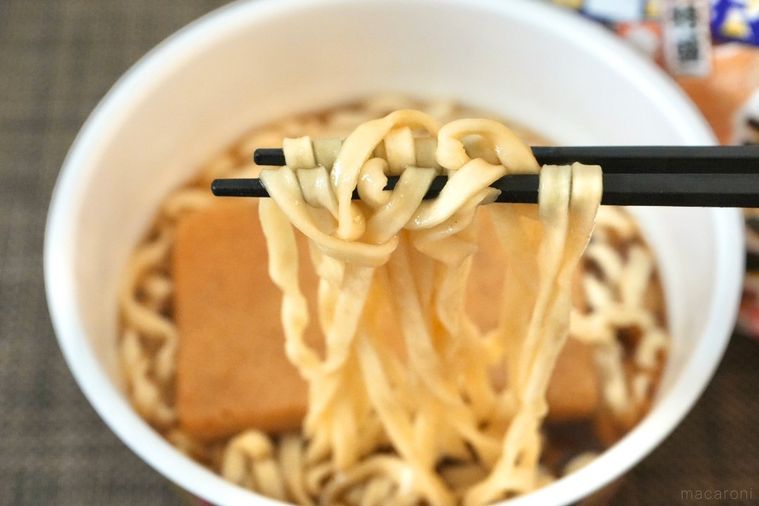 茶色いランチョンマットと大きなお揚げがのった煮干し味のカップうどんを黒い箸で食べる様子