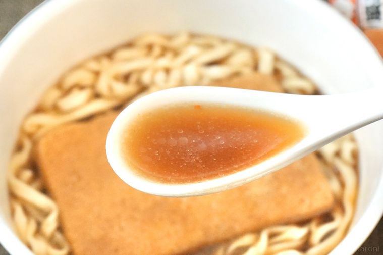 大きなお揚げがのった煮干し味のカップうどんのスープを白いレンゲですくう様子