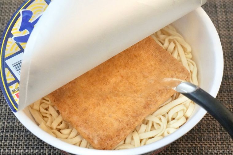 茶色いランチョンマットとフタを開けたカップうどんに黒い電子ケトルから熱湯を注ぐ様子