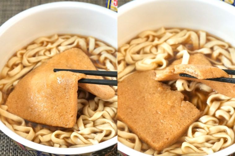 茶色いランチョンマットと煮干し味のカップうどんの大きなお揚げを黒い箸で持ち上げて割って食べる様子