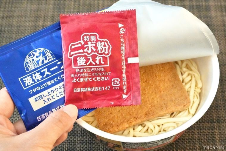 茶色いランチョンマットとフタを開けたカップきつねうどん煮干し味と味付けの粉末スープの赤色と青色の袋を手に持つ様子