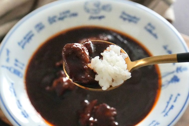 木のカッティングボードにのった白い器に盛られた牛タンビーフシチューと金と黒色のスプーンと生成色の布