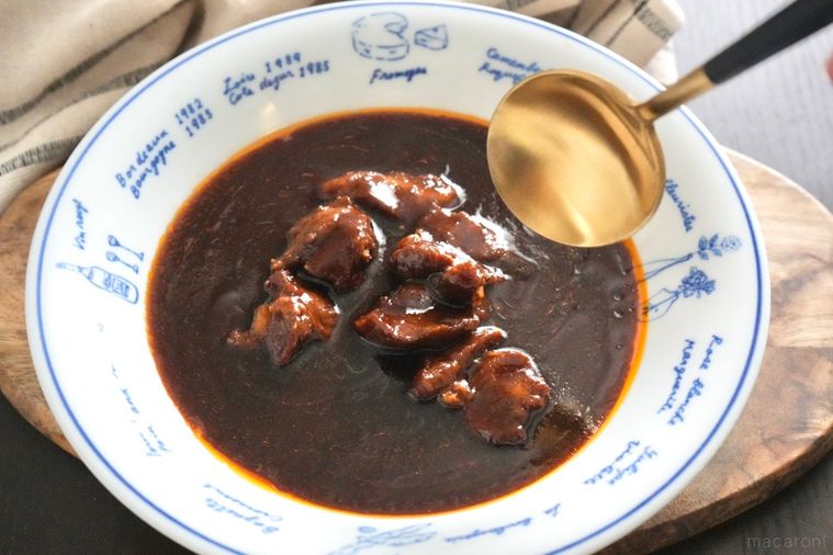 木のカッティングボードにのった白い器に盛られた牛タンビーフシチューと金と黒色のスプーンと生成色の布