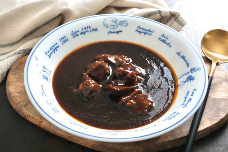 木のカッティングボードにのった白い器に盛られた牛タンビーフシチューと金と黒色のスプーンと生成色の布