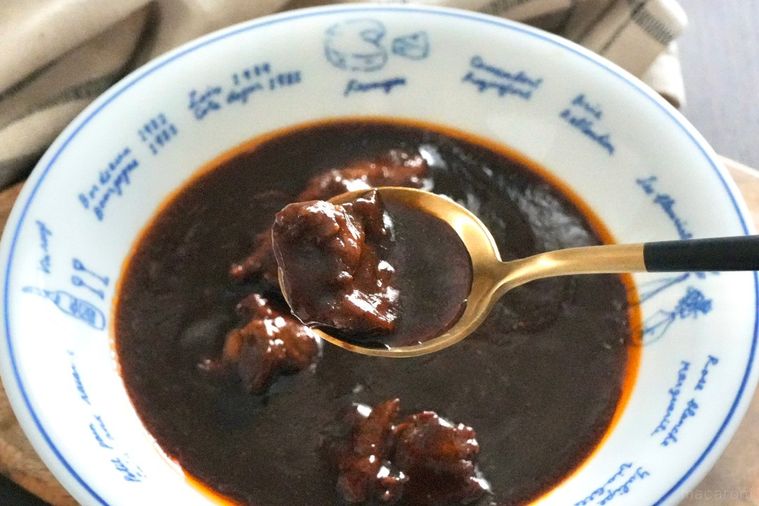 木のカッティングボードにのった白い器に盛られた牛タンビーフシチューと金と黒色のスプーンと生成色の布