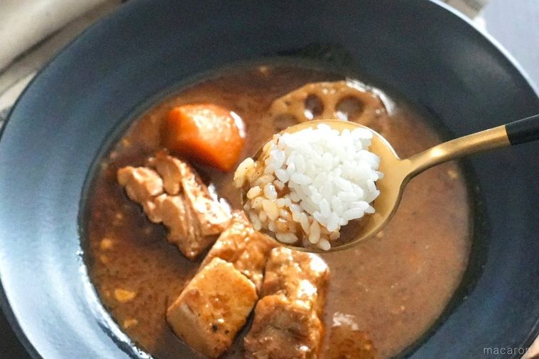 黒い皿に盛られたスープカレーを白ごはんと一緒に金と黒色のスプーンですくう様子と生成色の布
