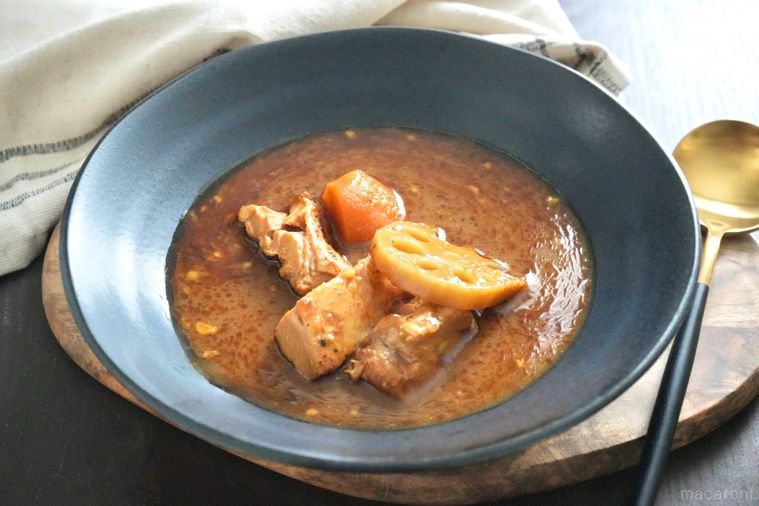 木のカッティングボードと黒い皿に盛られたにんじんとれんこんと鶏もも肉入りのスープカレーと金と黒色のスプーンと生成色の布