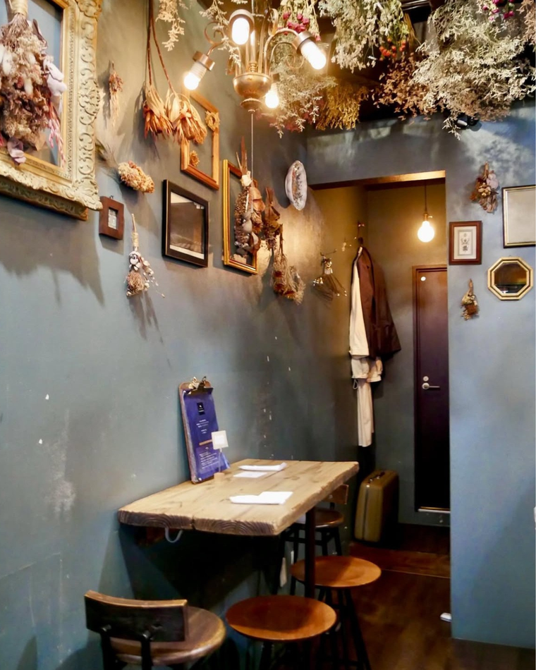 ドライフラワーやアンティーク額縁で彩られた壁と天井、木製のテーブルとスツールが並ぶ落ち着いたカフェの一角
