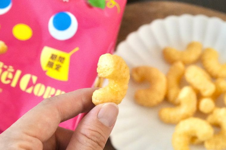 木のカッティングボードと白い皿にのった安納芋ブリュレ味のコーンスナックを持つ手とピンク色のパッケージ