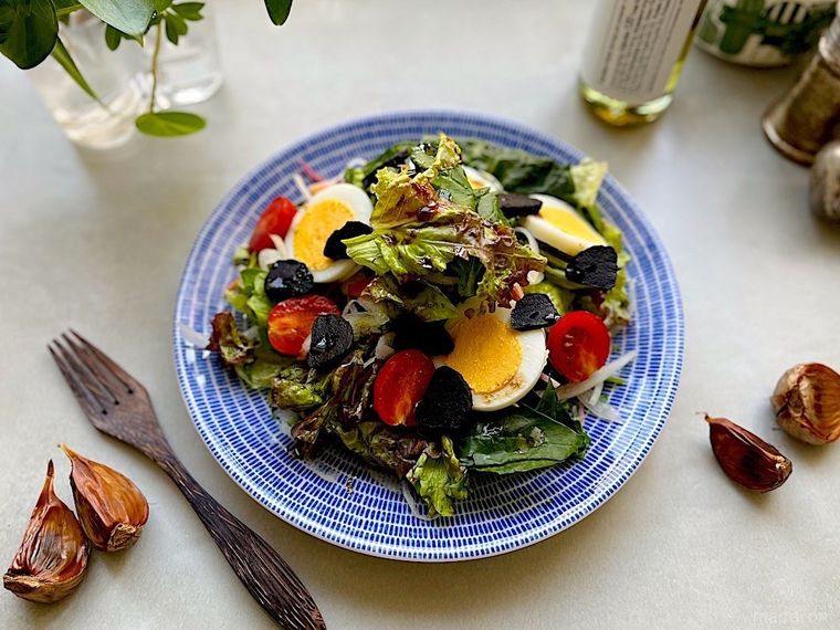 スライスしたコストコの黒にんにくを散らしたサラダ、皿のまわりに置かれた黒にんにく、カトラリー、調味料