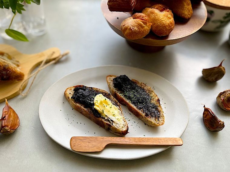 ペースト状にしたコストコの黒にんにくを塗った2切れのバゲットとカトラリー、皿のまわりに置かれた黒にんにく、いくつかの